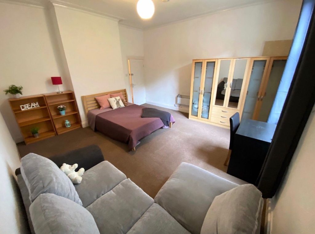 Bright bedroom with a double bed in mauve bedding, gray sofa in foreground, and a wooden shelves unit reading 'DREAM' on the left wall.